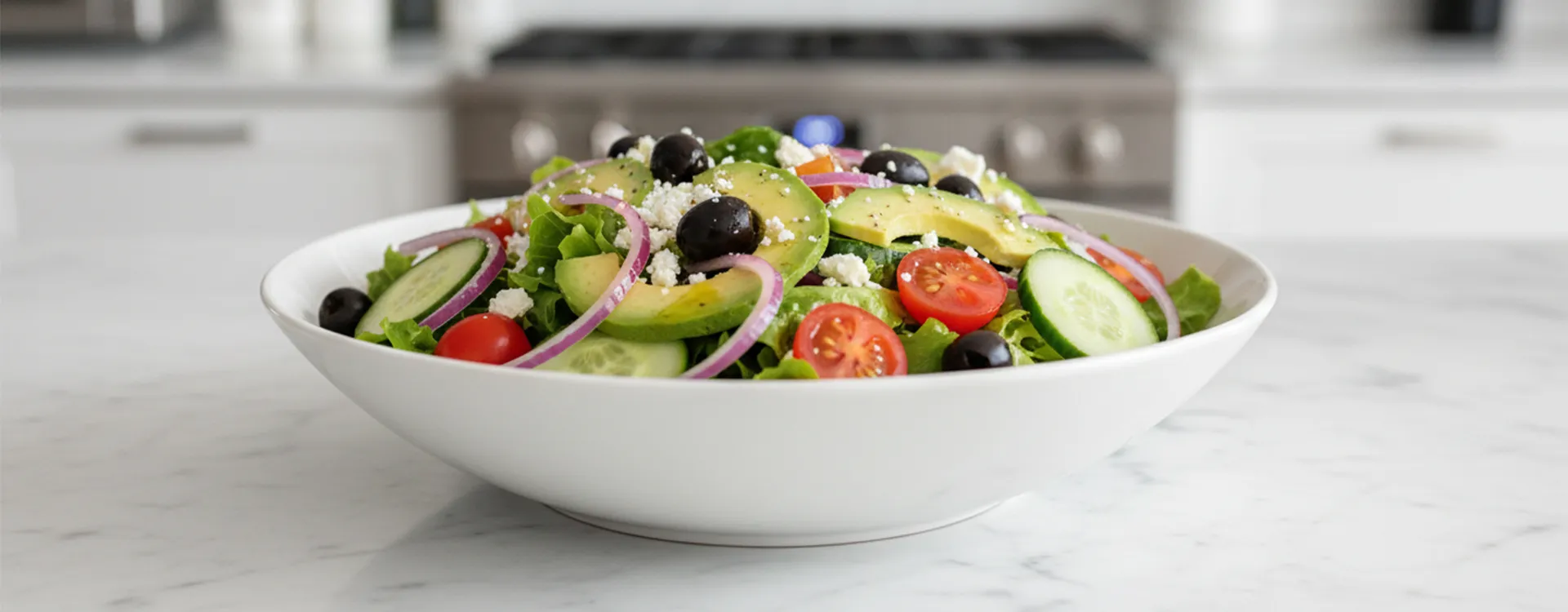 Large white bowl of fresh salad with lettuce, avocado, tomatoes, cucumbers, olives, onions, and feta on a marble kitchen counter.